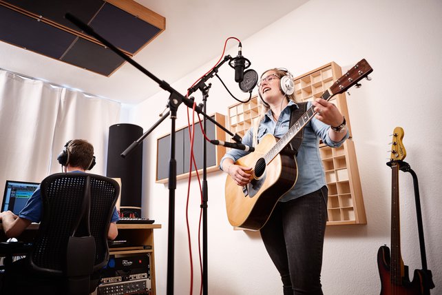 Eine junge Frau spielt Akustik-Gitarre und singt gleichzeitig. Sie trägt einen Studio-Kopfhörer und singt in ein Studio-Mikrofon, welches in einer Mikrofon-Spinne hängt. Im Hintergrund sitzt Florian Schmitz und bedient die Technik.